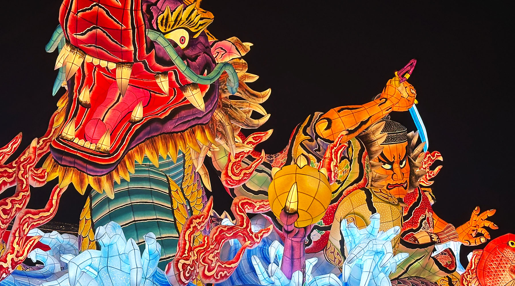 Aomori Nebuta Festival float featuring a powerful dragon and warrior illuminated against the night sky.
