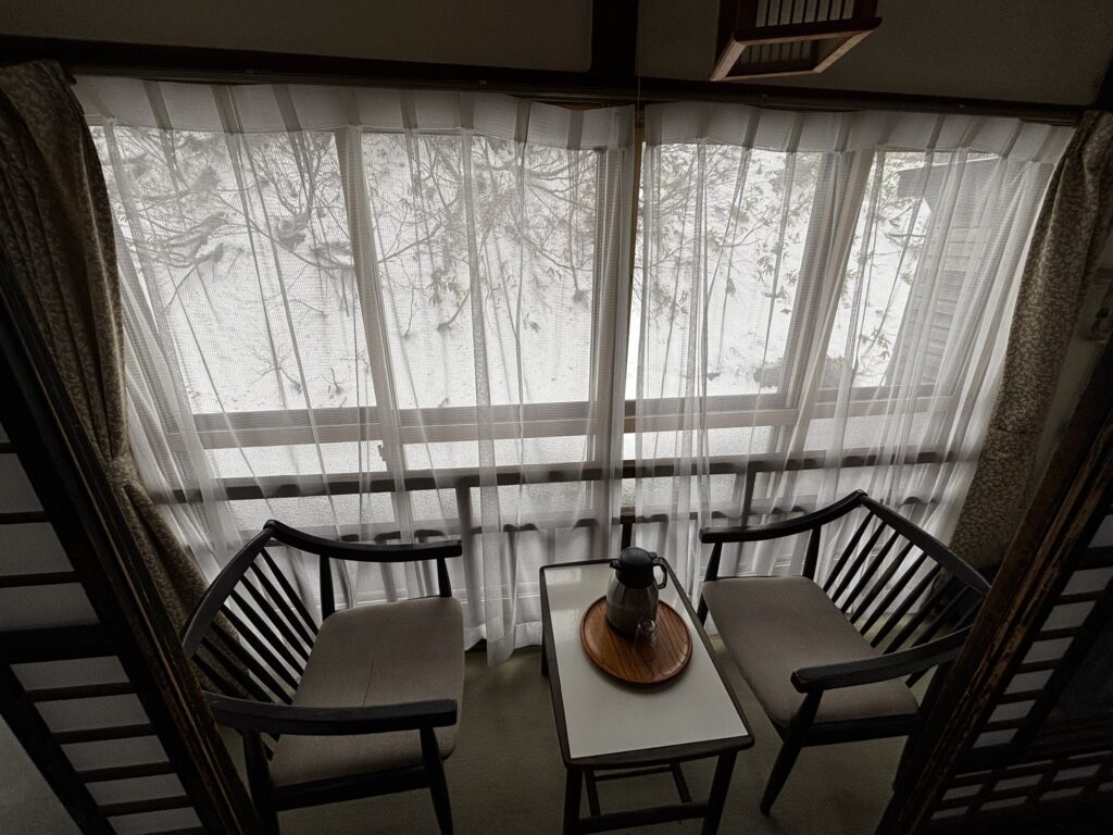 A hiroen sitting area at Sukayu Onsen with two chairs, a small table, and a snowy view through the lace curtains.